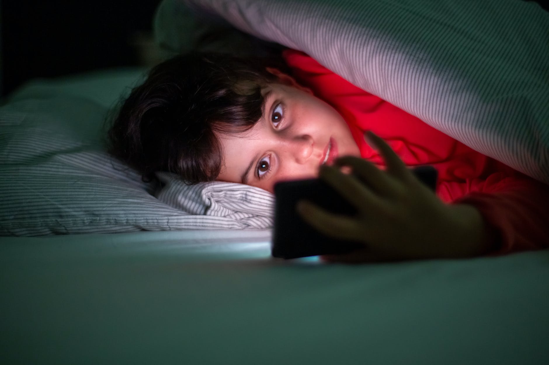 Kind ligt in bed onder het dekbed en kijkt op een smartphone